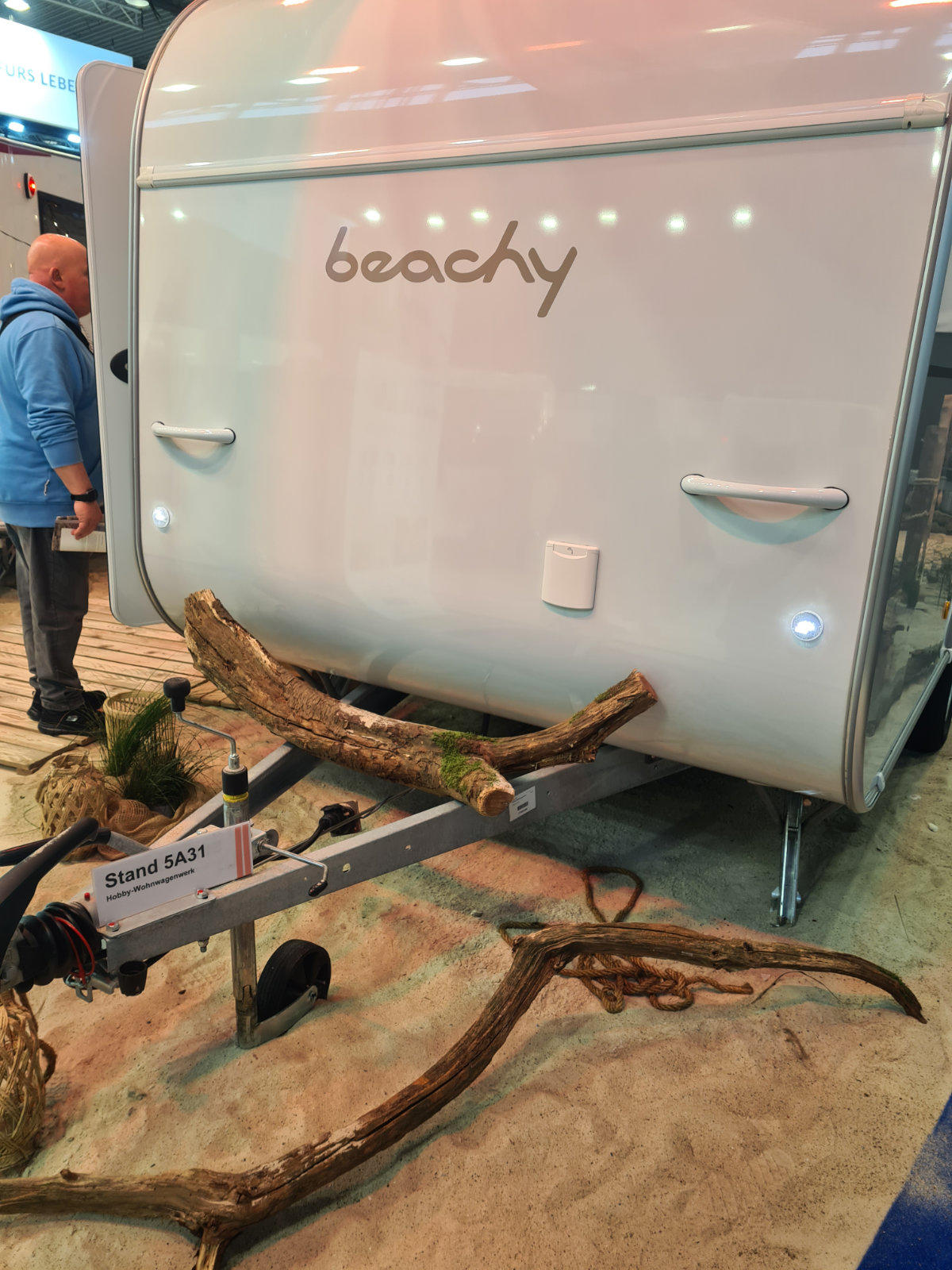 R&uuml;ckseite eines wei&szlig;en Wohnwagens mit Schriftzug 'beachy', Treibholzst&uuml;cke auf Sand davor, Anh&auml;ngerkupplung und links teilweise sichtbare Person