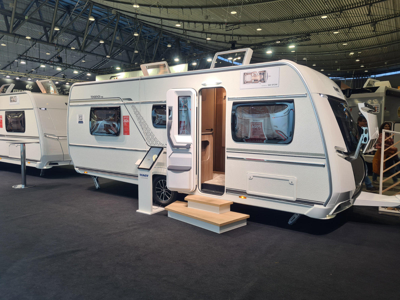 Wei&szlig;er Wohnwagen mit ge&ouml;ffneter T&uuml;r, einer Aufstiegsstufe davor, weitere Wohnwagen und hohe Hallendecke mit Beleuchtung im Hintergrund