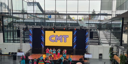 Gro&szlig;e Halle mit B&uuml;hne und gro&szlig;em Bildschirm, auf dem "CMT" steht; mehrere Personen in bunten Kost&uuml;men auf der B&uuml;hne; Publikum sitzt im Vordergrund; Glasfassade und Treppen im Hintergrund