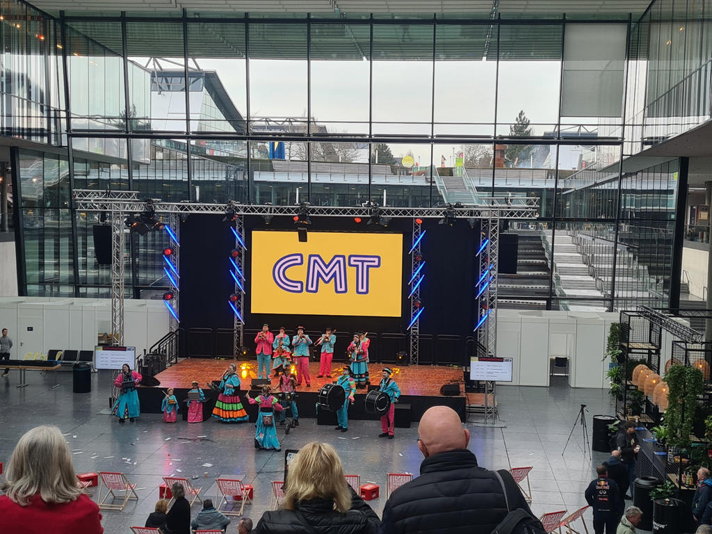 Gro&szlig;e Halle mit B&uuml;hne und gro&szlig;em Bildschirm, auf dem "CMT" steht; mehrere Personen in bunten Kost&uuml;men auf der B&uuml;hne; Publikum sitzt im Vordergrund; Glasfassade und Treppen im Hintergrund