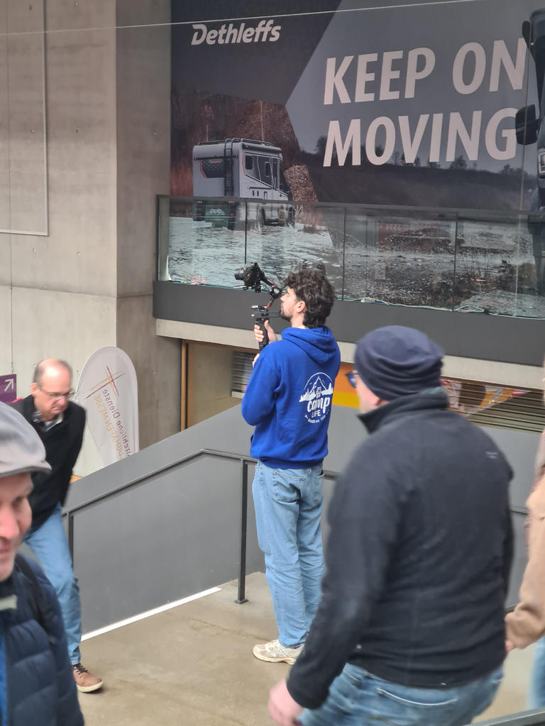 Person in blauem Kapuzenpullover h&auml;lt eine Kamera auf einem Stabilisator, steht auf einer Treppe; weitere Personen gehen vorbei; gro&szlig;es Werbeplakat mit Lkw und dem Schriftzug "KEEP ON MOVING" im Hintergrund