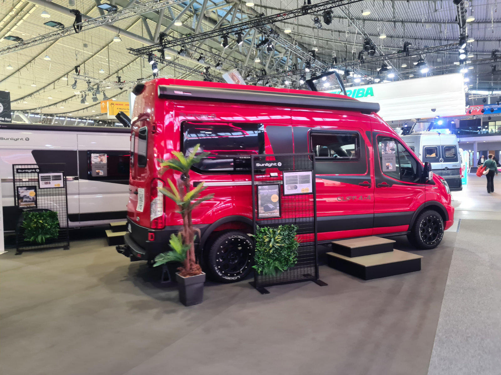 Rotes Campingfahrzeug in einer Messehalle, Seitenansicht mit schwarzen Felgen, Informationsst&auml;nder und Topfpflanzen daneben, kleine Treppe zum Einstieg, weitere Wohnmobile im Hintergrund