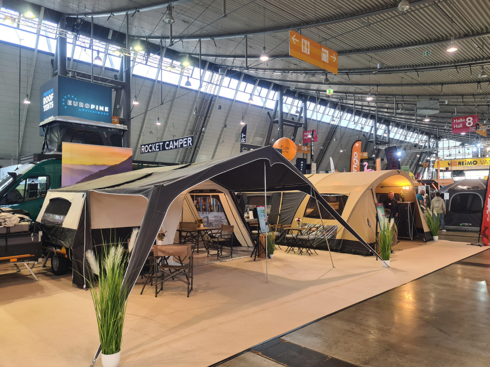 Gro&szlig;es Zelt mit Vordach auf einem Messestand in einer Ausstellungshalle, daneben Wohnwagen und Beschilderung "Rocket Camper"