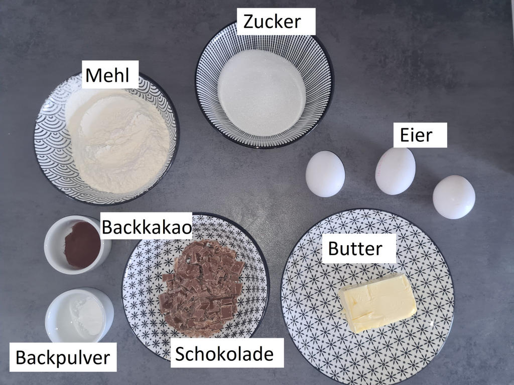 Sch&uuml;sseln und Teller mit Mehl, Zucker, Backkakao, Backpulver, Schokoladenst&uuml;cken, Butter; drei Eier auf grauem Untergrund