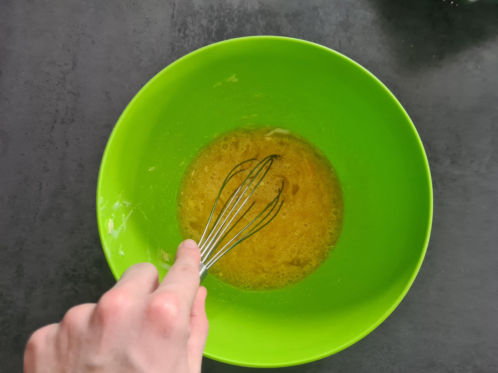 Gr&uuml;ne Sch&uuml;ssel mit gelblich-brauner Fl&uuml;ssigkeit, darin ein Metall-Schneebesen, an dem eine Hand den Griff h&auml;lt; dunkle Arbeitsfl&auml;che