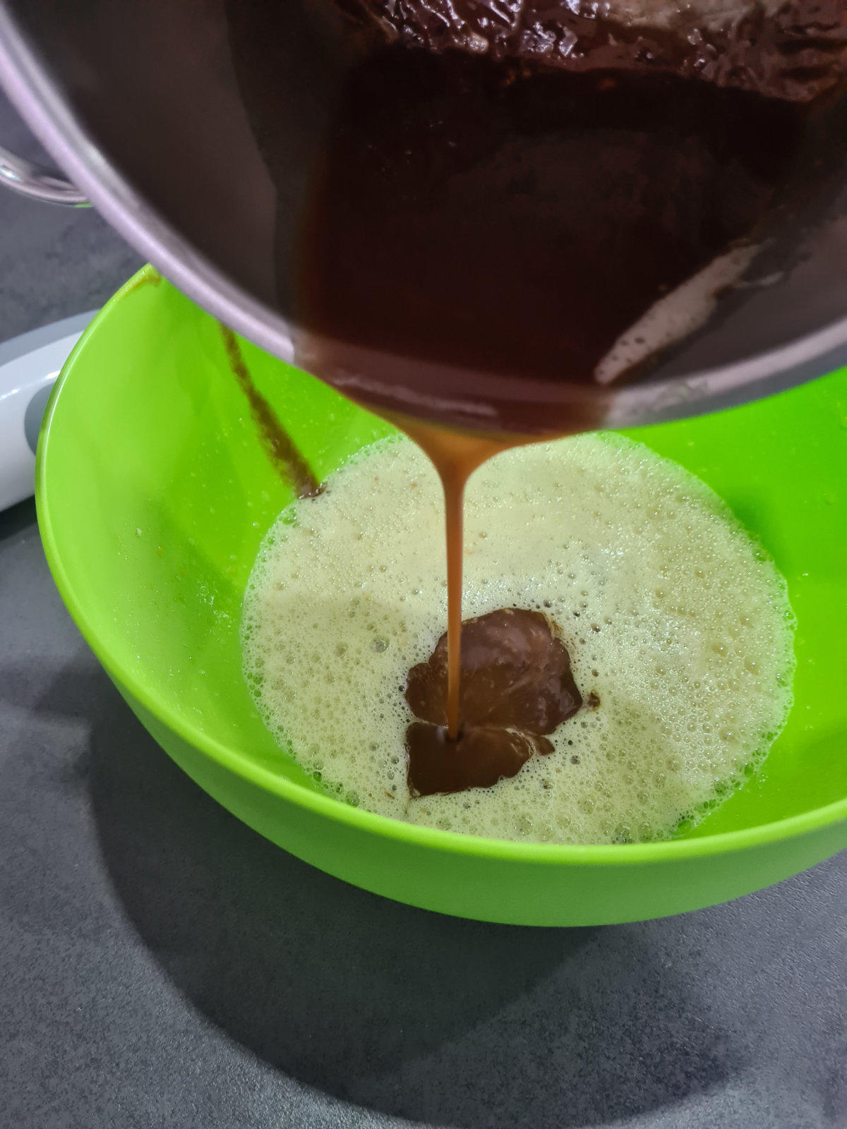 Geschmolzene Schokolade wird aus einem Metalltopf in eine gr&uuml;ne Sch&uuml;ssel mit schaumiger heller Fl&uuml;ssigkeit gegossen; wei&szlig;er Spatel im Topf