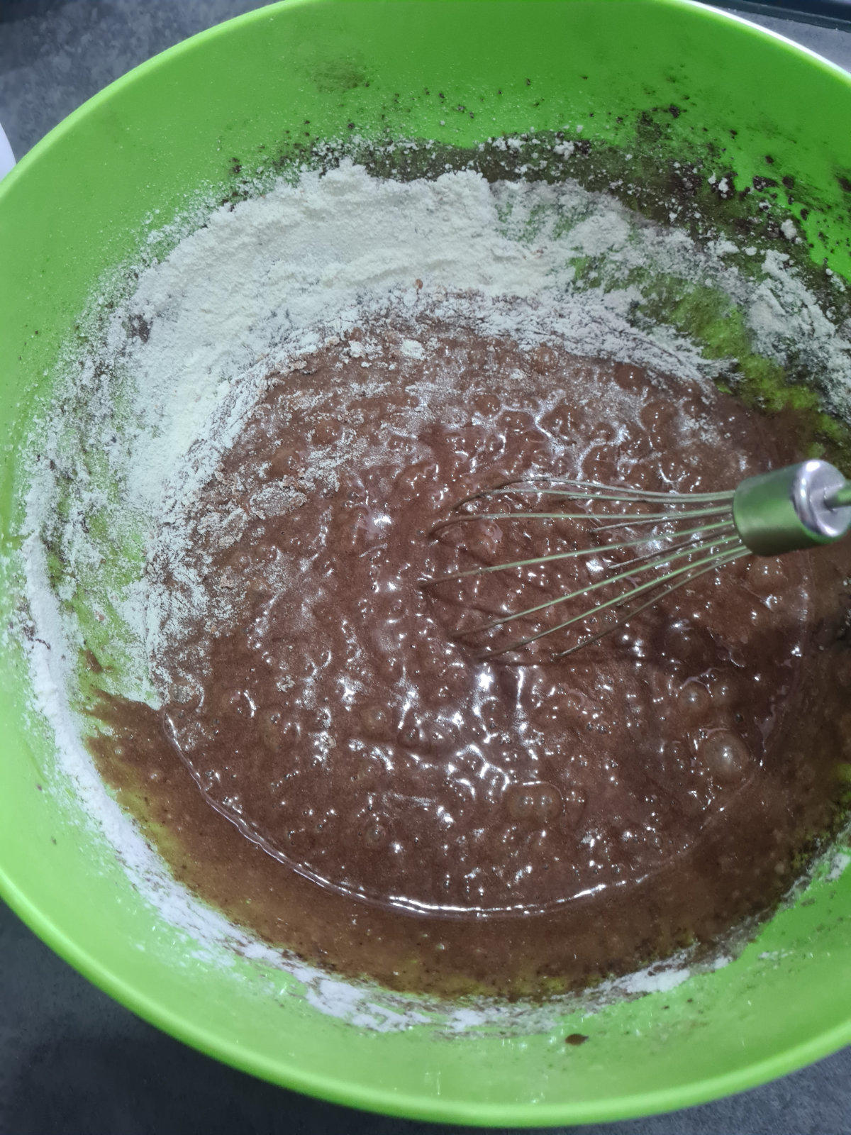 Gr&uuml;ne Sch&uuml;ssel mit wei&szlig;em Pulver am Rand und brauner gl&auml;nzender Masse in der Mitte; ein Schneebesen mit gr&uuml;nem Griff steckt darin; dunkle Arbeitsfl&auml;che im Hintergrund