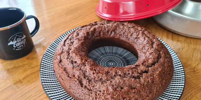Schokoladenkuchen in Ringform auf gemustertem Teller, roter Deckel einer Kuchenform und Metallform daneben, schwarze Tasse mit Logo auf Holztisch
