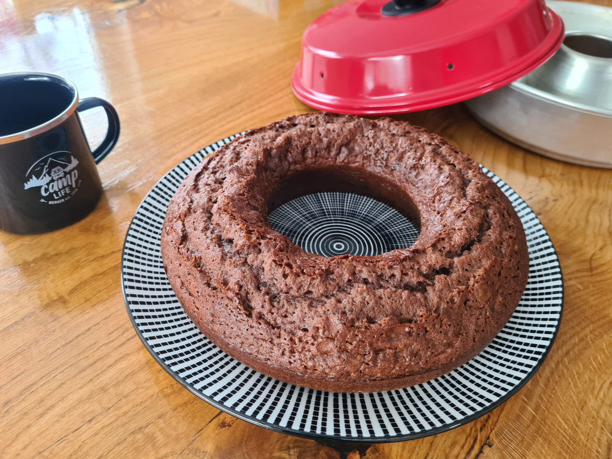 Schokoladenkuchen in Ringform auf gemustertem Teller, roter Deckel einer Kuchenform und Metallform daneben, schwarze Tasse mit Logo auf Holztisch