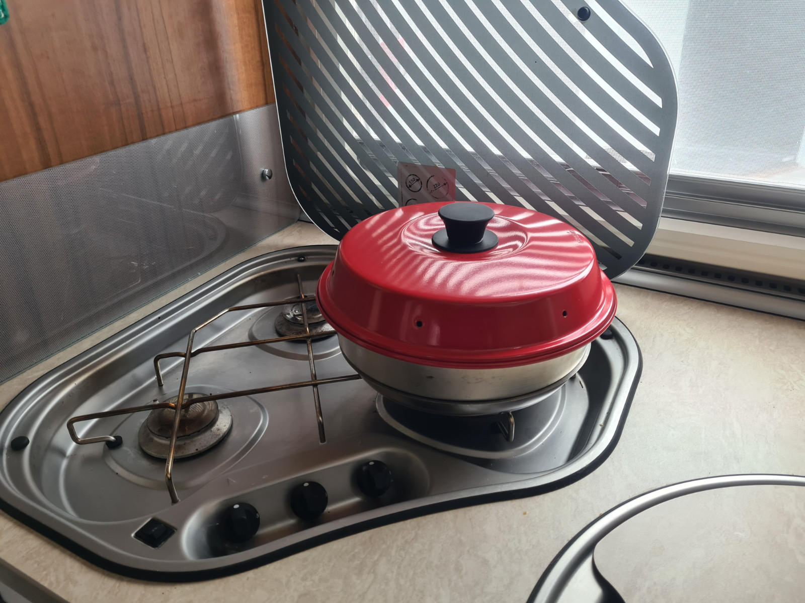 Runder roter Topf mit schwarzem Griff auf einem Gasbrenner; rechts perforierte Metallr&uuml;ckwand, zwei Brenner, drei schwarze Bedienkn&ouml;pfe, Holzpaneel, Fenster mit Jalousie, hellbeige Arbeitsfl&auml;che