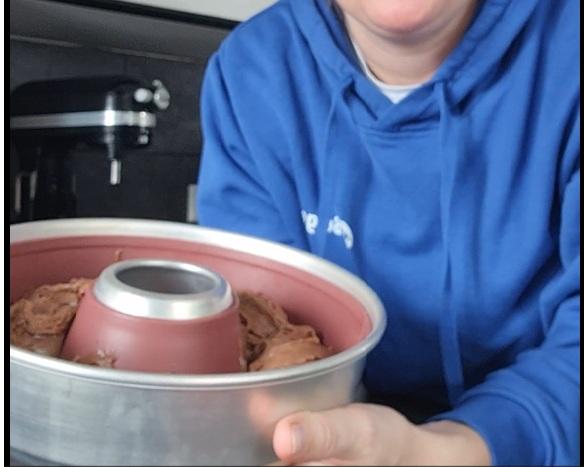 Person in blauem Kapuzenpullover, nur Untergesicht sichtbar, h&auml;lt eine runde Metall-Backform mit mittiger R&ouml;hre, innen rosa beschichtet, gef&uuml;llt mit brauner Masse