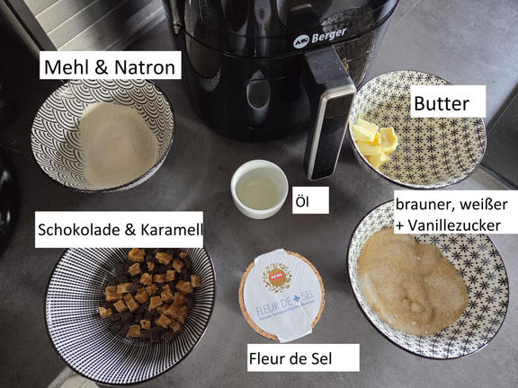 Plan de travail avec plusieurs bols : bol de poudre blanche &eacute;tiquet&eacute; 'Mehl & Natron', bol de sucre brun et blanc &eacute;tiquet&eacute; 'brauner, wei&szlig;er + Vanillezucker', morceaux de beurre, morceaux de chocolat et caramel, petit bol d'huile, bo&icirc;te 'Fleur de Sel', friteuse &agrave; air noire en arri&egrave;re‑plan