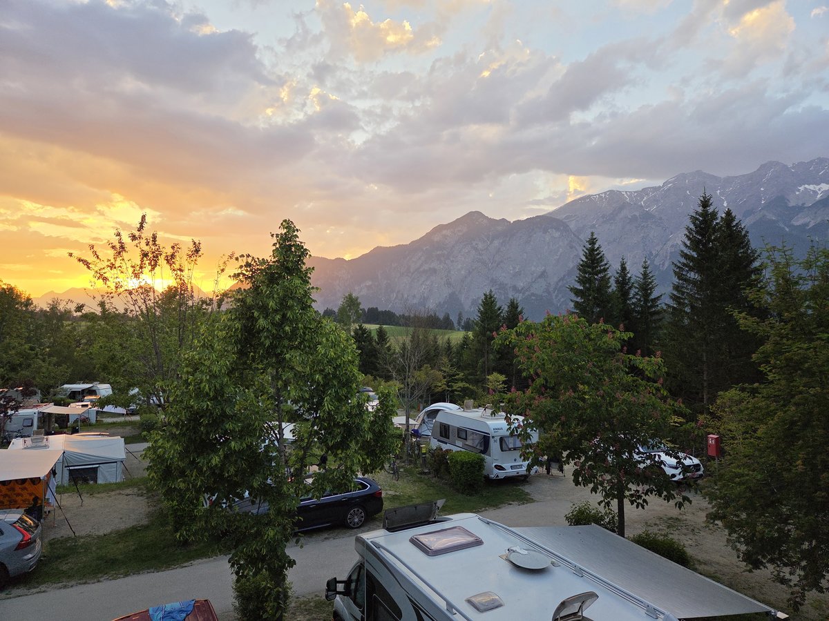 Campingplatz mit Wohnmobilen und Autos zwischen B&auml;umen, Berge im Hintergrund, orange-blauer bew&ouml;lkter Himmel