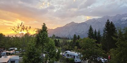 Campingplatz mit Wohnmobilen und Autos zwischen B&auml;umen, Berge im Hintergrund, orange-blauer bew&ouml;lkter Himmel