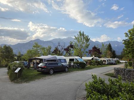 Mehrere geparkte Wohnmobile und Autos auf einer grasigen Stellfl&auml;che neben einem Asphaltweg, B&auml;ume, Berge im Hintergrund und wolkiger Himmel