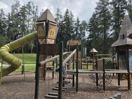 Holzspielplatz mit T&uuml;rmen, Rutschen, Seilbr&uuml;cken und Kletterger&uuml;sten vor hohen Nadelb&auml;umen unter bew&ouml;lktem Himmel