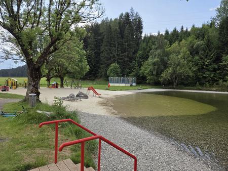 Badeweiher mit flachem Ufer aus Kies und Sand, rotes Gel&auml;nder an Stufen im Vordergrund, Spielger&auml;te und B&auml;ume, Wald im Hintergrund