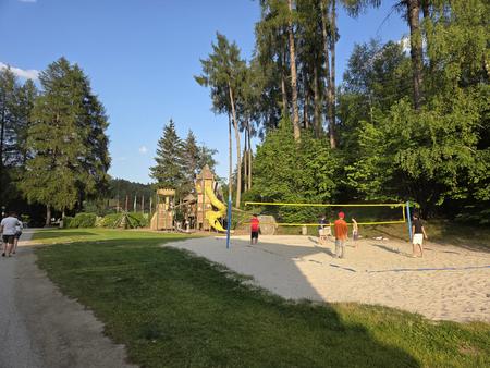 Sand-Volleyballfeld mit mehreren Personen, Volleyballnetz rechts, Spielplatz mit gelber Rutsche links, hohe Nadelb&auml;ume im Hintergrund und blauer Himmel