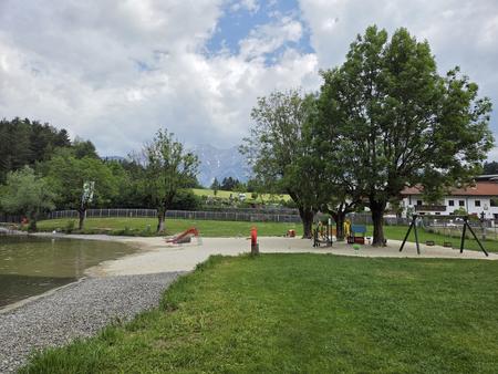 Ufer eines kleinen Sees mit Sandstrand, einer Rutsche und einer Schaukel, B&auml;umen, gr&uuml;ner Wiese, H&auml;usern im Hintergrund und bew&ouml;lktem Himmel