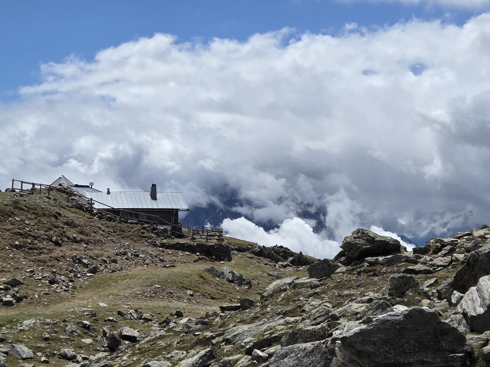 Steiniger Berghang mit Grasfl&auml;chen und Felsen, links ein Geb&auml;ude mit Metalldach, im Hintergrund tiefe Wolken und teils sichtbare Berggipfel