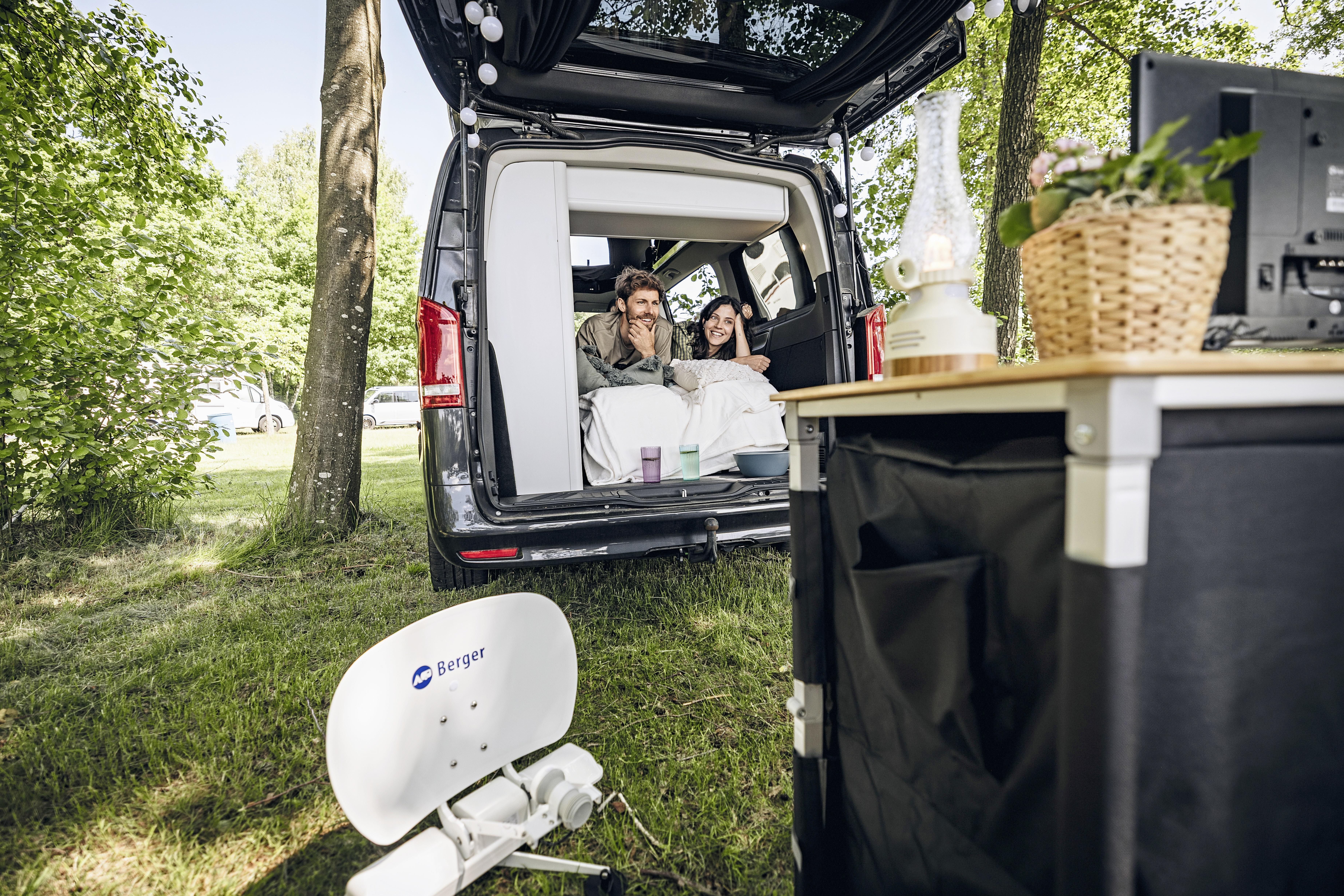 Zwei Personen sitzen im ge&ouml;ffneten Laderaum eines Vans; davor auf dem Gras eine tragbare Satellitensch&uuml;ssel und ein Tisch mit einem Pflanzenkorb