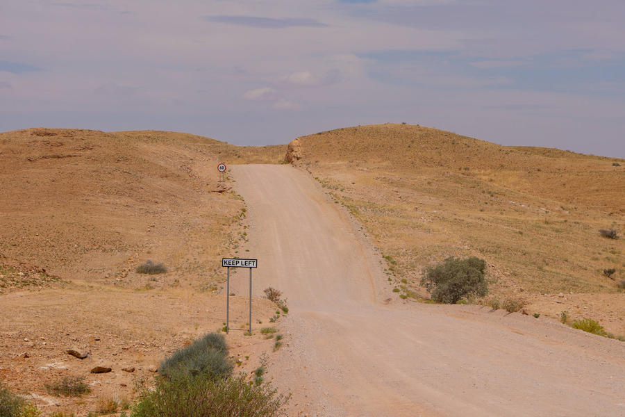 Unbefestigte Schotterstra&szlig;e zwischen niedrigen sandigen H&uuml;geln in einer trockenen Landschaft, kleine Str&auml;ucher am Stra&szlig;enrand, Metallwegweiser, hellblauer Himmel mit d&uuml;nnen Wolken