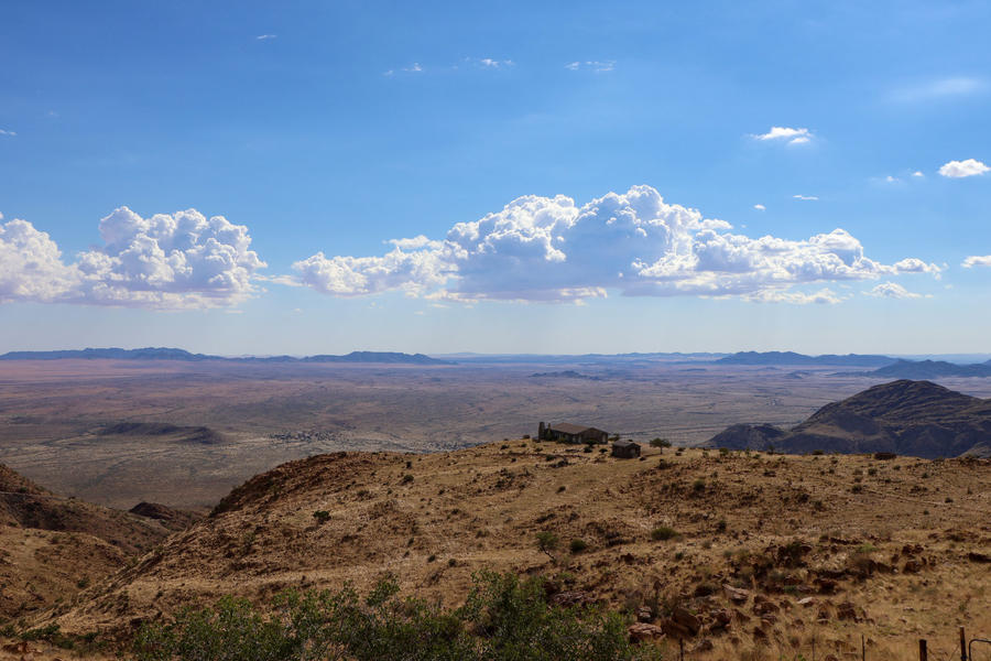 Felsiger, brauner Vordergrund mit vereinzelten gr&uuml;nen Pflanzen; dahinter eine weite Ebene mit flachen H&uuml;geln und niedrigen Tafelbergen am Horizont; blauer Himmel mit verstreuten wei&szlig;en Wolken