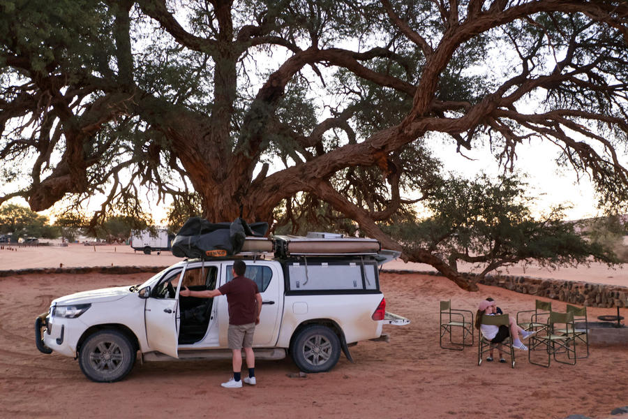 Wei&szlig;er Pickup mit Gep&auml;ck auf dem Dach und offener Fahrert&uuml;r vor einem gro&szlig;en knorrigen Baum auf rotem Sand; eine Person steht neben dem Fahrzeug, zwei Personen sitzen rechts an einem Campingtisch mit St&uuml;hlen