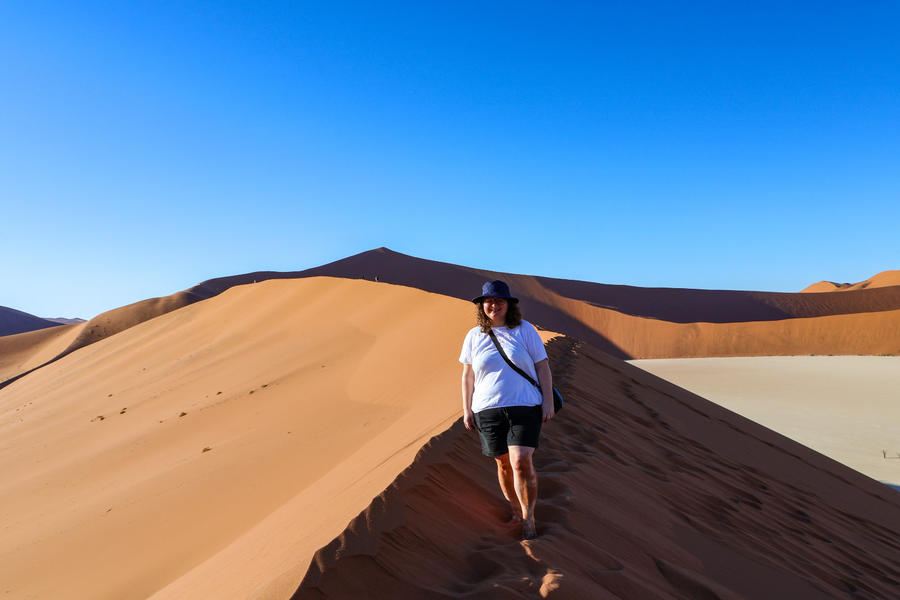 Eine Person in wei&szlig;em T-Shirt, schwarzen Shorts und dunklem Hut geht barfu&szlig; auf dem Grat einer orangefarbenen Sandd&uuml;ne; Fu&szlig;spuren im Sand, klarer blauer Himmel