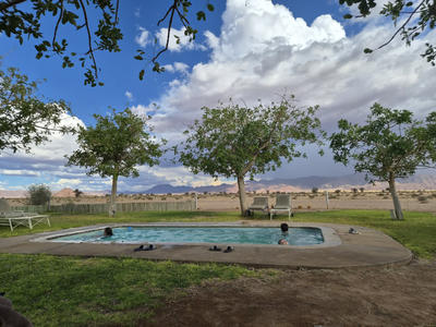 Rechteckiges Au&szlig;enschwimmbecken in einem Garten mit mehreren Personen im Wasser, Rasen, drei B&auml;ume, eine Bank und Schuhe am Beckenrand, Zaun, Berge im Hintergrund und wolkiger Himmel