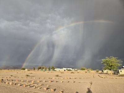 Doppelter Regenbogen &uuml;ber sandiger Fl&auml;che mit Reihen kleiner Erdh&uuml;gel, dunklen Regenwolken und einem Baum rechts, im Vordergrund Schatten einer Person