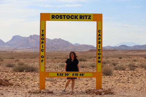 Person steht in einem gro&szlig;en gelben Quadratrahmen mit der Aufschrift 'ROSTOCK RITZ', 'TROPIC OF' und 'CAPRICORN' und den Koordinaten S 23&deg; 30' E 15&deg; 46'; steiniger W&uuml;stenboden, Berge am Horizont und heller Himmel