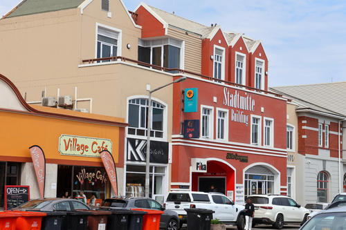 Stra&szlig;e mit bunten Gesch&auml;ftsgeb&auml;uden, orange Caf&eacute; mit Schild &bdquo;Village Caf&eacute;&ldquo;, rotes Geb&auml;ude mit wei&szlig;er Schrift, geparkte Autos und M&uuml;lltonnen, blauer Himmel