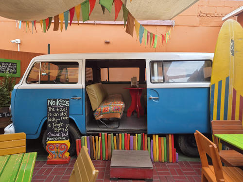Blauer und wei&szlig;er Bus mit ge&ouml;ffneter Schiebet&uuml;r, Kissen und kleinem Tisch im Inneren; bunte Wimpel oben; gelbes Surfbrett rechts; bunte Holzlatten vor dem Bus; bemalte Tische und St&uuml;hle im Vordergrund; Kreidetafel links