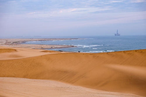 Sandd&uuml;nen im Vordergrund, K&uuml;stenlinie mit Wellen und eine Plattform im Meer in der Ferne