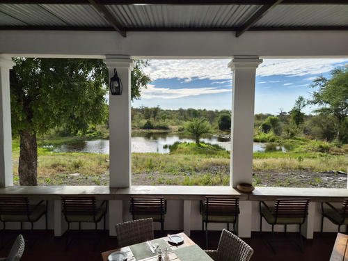 &Uuml;berdachte Veranda mit drei wei&szlig;en S&auml;ulen, Tisch mit Besteck und Gl&auml;sern im Vordergrund, Reihe von St&uuml;hlen an der Br&uuml;stung, Teich mit Ufervegetation, B&auml;ume und wolkiger Himmel
