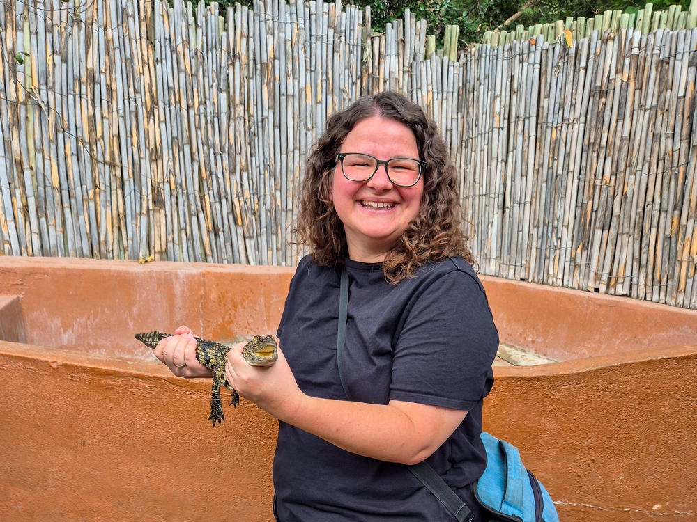 Person mit lockigen schulterlangen Haaren und Brille h&auml;lt eine kleine Schildkr&ouml;te vor einer Bambuswand und einer niedrigen terrakottafarbenen Mauer