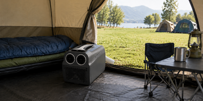 Blick aus einem ge&ouml;ffneten Zelt: Schlafsack auf Feldbett, tragbares Ger&auml;t mit L&uuml;ftungsschlitzen auf dem Boden, Klappstuhl und Campingtisch mit Metallbecher und Laterne; drau&szlig;en Wiese, weitere Zelte, B&auml;ume, See und Berge
