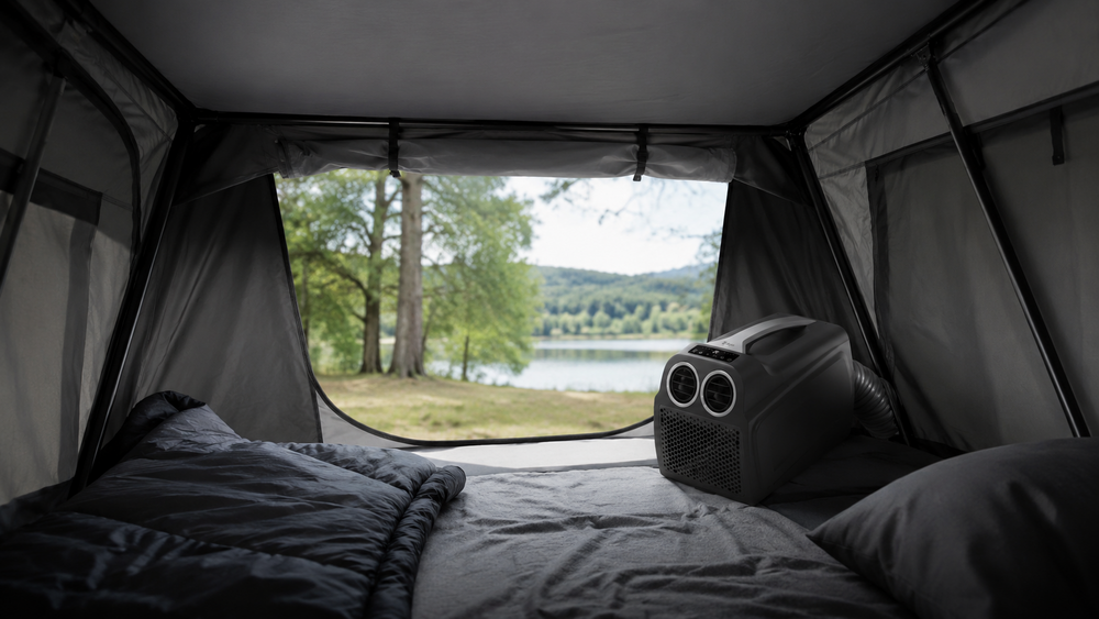 Blick aus einem Zelt auf einen See und eine baumbewachsene Uferlinie; im Zelt liegen ein Schlafsack, ein Kissen und ein tragbares Gerät mit zwei runden Lüftungsgittern