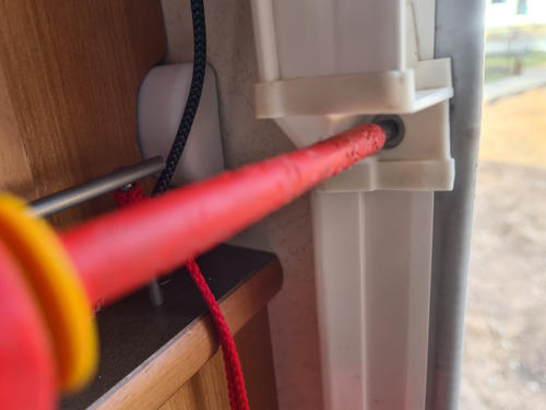 Roter Schraubendreher steckt in wei&szlig;em Kunststoffhalter an einem Rahmen; links Holzblende, schwarze Kabel und rotes Textilkabel auf einer Ablage, rechts unscharfer Au&szlig;enbereich