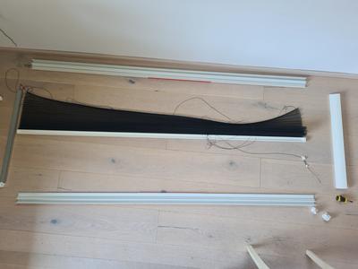 Heller Holzboden mit zwei langen wei&szlig;en Metallprofilen parallel liegend, ein schwarzes Stoffst&uuml;ck h&auml;ngt zwischen ihnen, mehrere d&uuml;nne schwarze Kabel und kleine wei&szlig;e und gelbe Kunststoffteile, wei&szlig;e Wand am oberen Bildrand