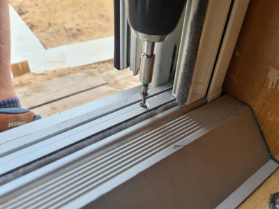 Bohrmaschine mit Schraubbit &uuml;ber einer Schraube in einer Aluminium-T&uuml;rschwelle, Dichtung am Rahmen, Holzboden und Fu&szlig; mit Sandale im Hintergrund