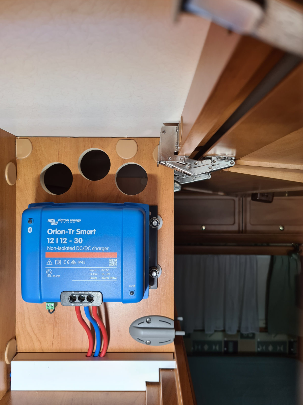 Blaues Ger&auml;t mit Aufschrift 'Orion-Tr Smart 12 | 12-30' an einer Holzplatte montiert, drei runde &Ouml;ffnungen dar&uuml;ber, drei Kabel (rot, blau, schwarz) f&uuml;hren nach unten, rechts ein Metallscharnier, im Hintergrund Fenster mit Vorhang