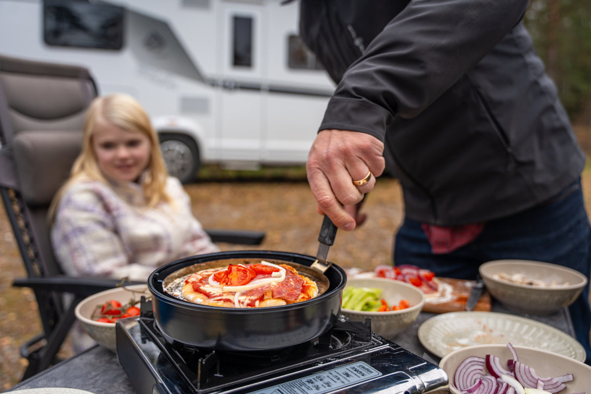 Hand einer erwachsenen Person r&uuml;hrt mit einem Spatel in einer Pfanne auf einem Campingkocher; auf dem Tisch Sch&uuml;sseln mit Salat, Tomaten und Zwiebeln; im Hintergrund sitzt ein M&auml;dchen auf einem Campingstuhl, dahinter ein Wohnmobil