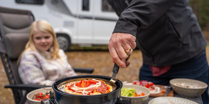 Hand einer erwachsenen Person r&uuml;hrt mit einem Spatel in einer Pfanne auf einem Campingkocher; auf dem Tisch Sch&uuml;sseln mit Salat, Tomaten und Zwiebeln; im Hintergrund sitzt ein M&auml;dchen auf einem Campingstuhl, dahinter ein Wohnmobil