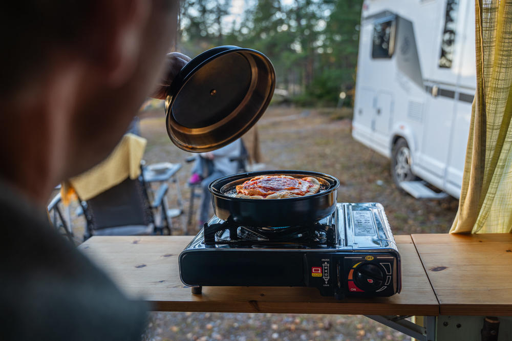 Person hebt Topfdeckel &uuml;ber eine Pfanne mit Pizza auf einem tragbaren Gaskocher auf einem Holztisch, Wohnmobil und B&auml;ume im Hintergrund