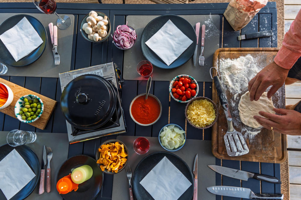 Tisch mit Tellern, Servietten und Besteck; Sch&uuml;sseln mit Tomatenso&szlig;e, geriebenem K&auml;se, geschnittener Zwiebel, Kirschtomaten, Oliven, Pilzen und geschnittenen Paprika; Holzbrett mit Mehl und H&auml;nden, die Teig formen; Pfanne auf tragbarem Herd