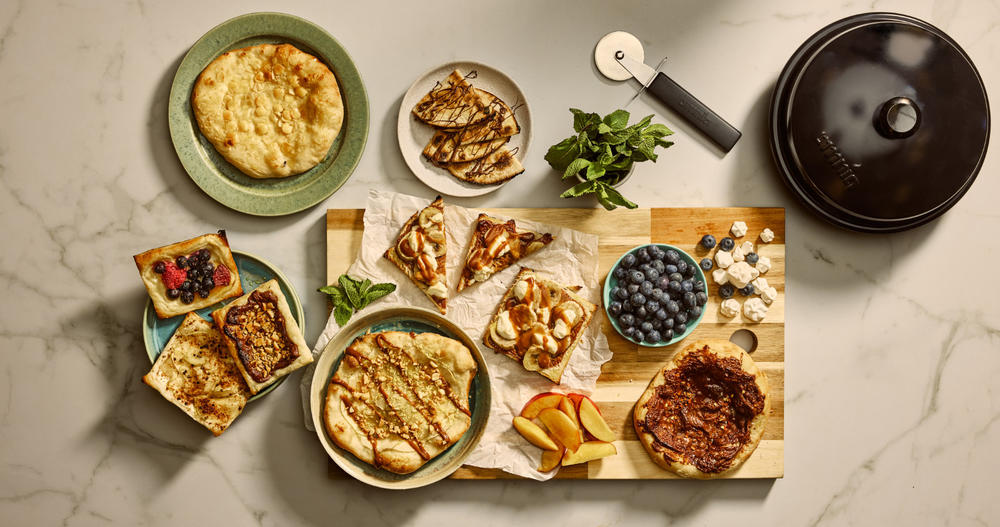 Holzbrett auf Marmor mit gefalteten d&uuml;nnen Teigfladen mit Grillstreifen, runden Pfannkuchen, Toastscheiben mit Beerenaufstrich, Schale mit Blaubeeren, Sch&uuml;ssel mit dunkler Creme, Apfelscheiben, kleine wei&szlig;e St&uuml;cke, Minzblatt, Pizzaschneider und Pfann