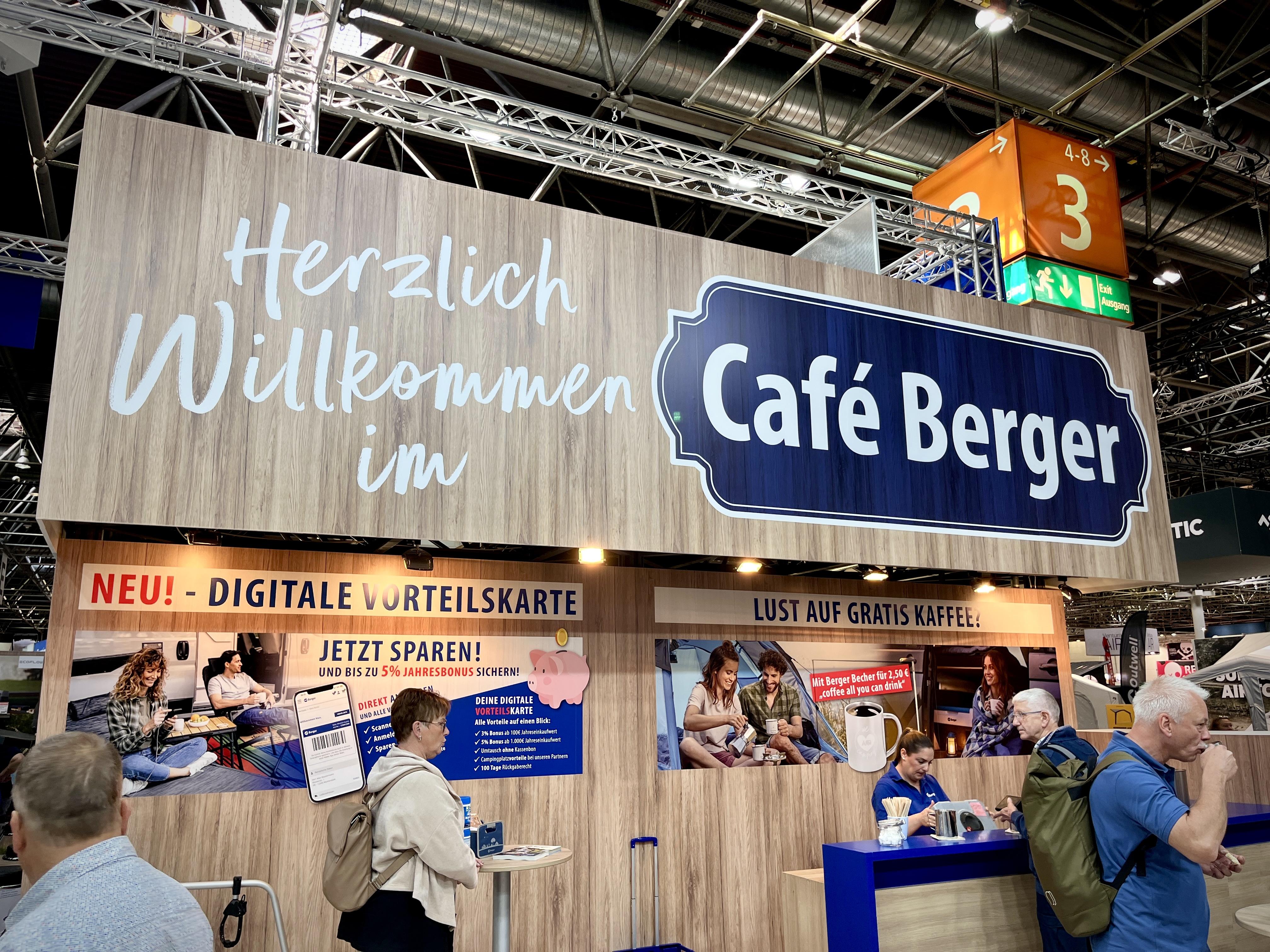 Messestand mit Holzverkleidung und gro&szlig;em Schild mit der Aufschrift 'Herzlich Willkommen im Caf&eacute; Berger', Werbebanner 'NEU! - DIGITALE VORTEILSKARTE', mehrere Personen davor, orangefarbiges Hallenschild mit '3'