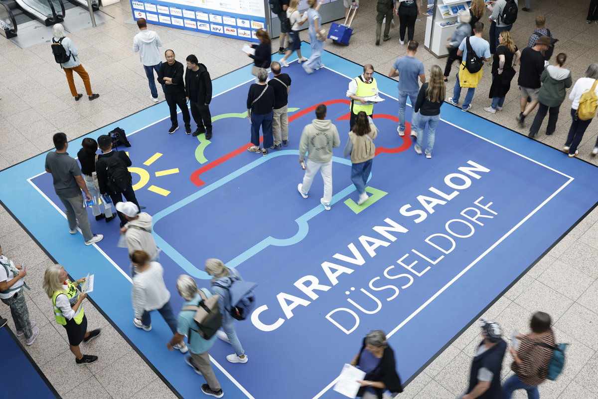 Aufnahme von oben: blauer Bodenaufkleber mit Schrift 'CARAVAN SALON D&Uuml;SSELDORF', stilisierte Wohnwagen-Grafik und mehrere Personen in einer Halle